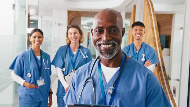 Close up portrait of smiling mature male doctor or nurse with digital tablet wearing scrubs standing in hospital corridor with multi-cultural team of colleagues in background - shot in real time