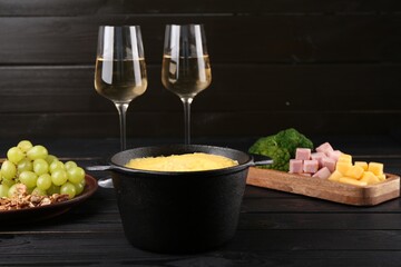 Fondue pot with melted cheese, glasses of wine and different products on black wooden table