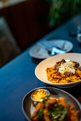 Eggplant pasta, onsen eggs, spiced chicken wings, fried food, restaurant food, casual entertainment, wooden tabletop, background, white ceramic plate, metal saucer, dipping sauce