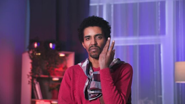 young black man touching his face with his hands and looking thoughtfully at camera while standing in room