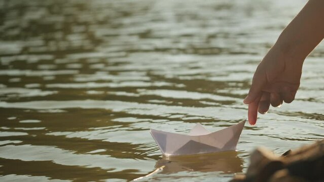 Woman hand puts paper ship boat on water in the park close up. Origami paper crane on a pond. Natural lake view on sunny day