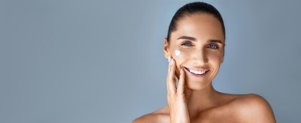 Anti-aging cosmetics. Portrait of beautiful mature woman applaing face skin care product ,smiling, posing against grey background