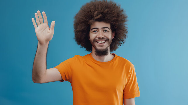 Portrait of cheerful friendly man with Afro hairstyle wearing orange T-shirt saying hi and waving hand, greeting, looking at camera. Indoor studio shot isolated on blue background.