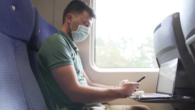 Man working in train, wearing masks to prevent air pollution, dust, PM2.5 pm2, coronavirus or COVID 19. Male passenger is surfing internet on smart phone and uses laptop near window. Railway transport