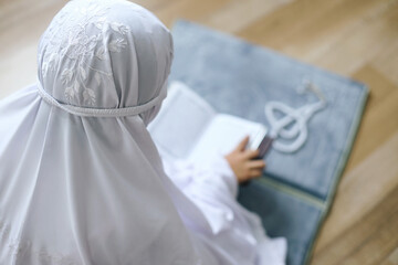 Rear view of young muslim girl reading Quran on prayer mat