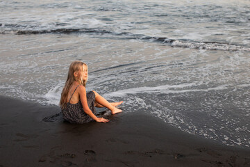 beautiful teenage girl sitting on the ocean shore and dreaming, black volcanic sand, volcanic sand beach