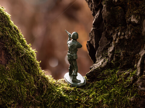 A toy soldier stands on a green moss covered brown tree branch, enduring the scorching heat of a hot summer heatwave weather. In the background, nature is blurred.