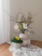 A vase of flowers and a homemade primrose flower in a pot on a round wooden table with a white tablecloth