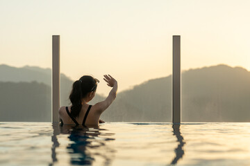 Beautiful white woman wearing a black bikini relaxing in swimming pool roof top at luxury hotel spa enjoying beautiful golden sunset view of mountains, nature and city.