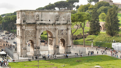 The Arch of Constantine near the Colosseum, under a cloudy sky, surrounded by tourists and green...