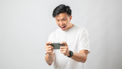 Fototapeta premium Portrait of an Indonesian Asian man, wearing a white T-shirt, expressively playing a game on his smartphone, isolated against a white background.