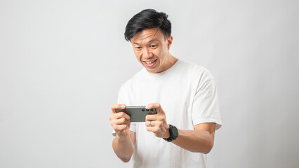 Portrait of an Indonesian Asian man, wearing a white T-shirt, expressively playing a game on his smartphone, isolated against a white background.