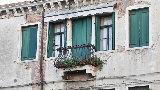An aged building facade with a single balcony filled with plants and green shutters, showing weathered textures and a sense of history.