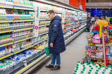 male man in winter clothes chooses sour cream in dairy section of local market, store. Supermarket open refrigerator, wide selection of dairy products. Pavlodar, Kazakhstan 1.17.23