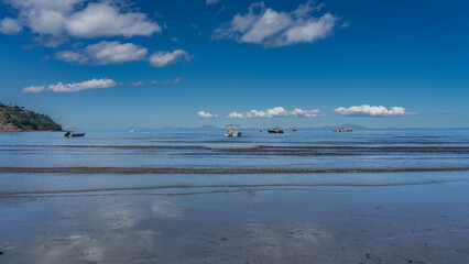 Boats are visible in the blue ocean. the waves roll towards the shore and spread out on the beach. Mountains on the horizon. Clouds in the azure sky. Madagascar. Nosy Be  