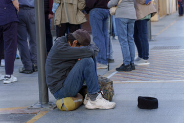 homeless man on the street begging for alms with a sad expression, his head between his legs and a...