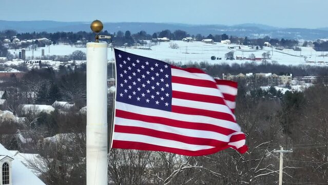 American flag waving in front of snow covered winter landscape in town. Aerial with zoom lens.