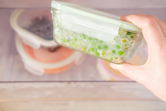 Female hand holds a glass container with green peas against the background of freezer with other boxes.