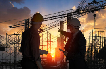 Two construction engineers in hard hats discuss project plans against a backdrop of silhouetted cranes at sunset..