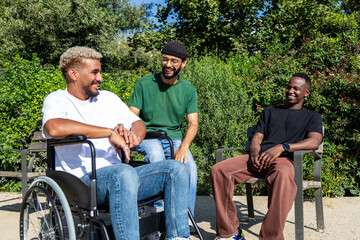 Young disabled African American man in a wheelchair and his male friends talking sitting on a bench in city park.