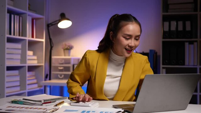 Cheerful Asian businesswoman using a laptop and writing on the charger to take notes. Analyzing finances, online markets, and taxes and happily collecting thoughts and ideas in her office.