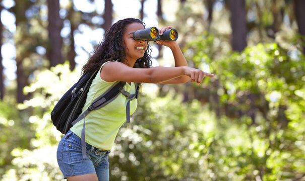 Happy Girl, Binoculars And Pointing In Nature With Backpack For Sightseeing, Explore Or Outdoor Vision. Young Female Person, Child Or Teenager Enjoying Adventure, Bird Watching Or Search In Forest