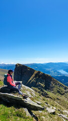 Naklejka premium Hiker woman sitting on rock on alpine meadow with panoramic view of lake Millstatt. Idyllic hiking trail to mountain peak Boese Nase, Ankogel Group, Carinthia, Austria. Remote Austrian Alps in summer