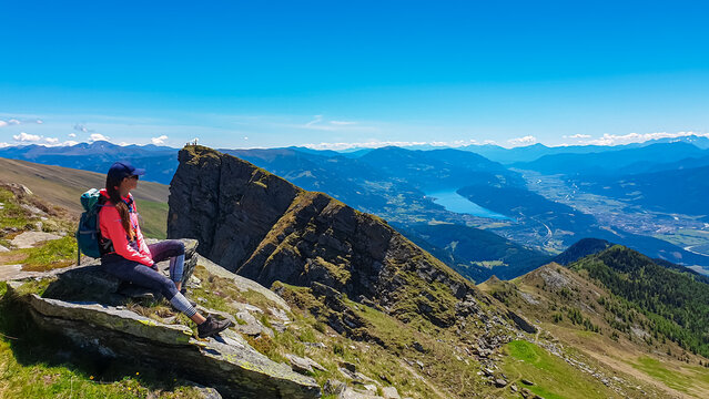 Hiker woman sitting on rock on alpine meadow with panoramic view of lake Millstatt. Idyllic hiking trail to mountain peak Boese Nase, Ankogel Group, Carinthia, Austria. Remote Austrian Alps in summer