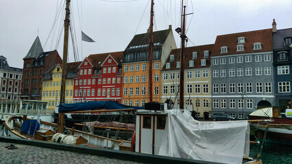 Historic European buildings with gabled roofs line a harbor with docked boats and masts rising...