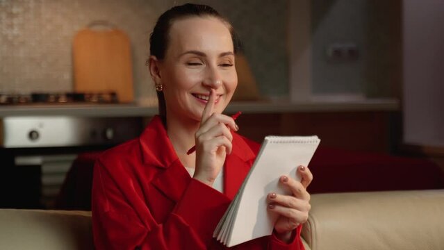 A woman dressed in a vibrant red blaser thoughtfully touched her face with a pen while holding a notepad, looking away in a warmly lit indoor setting.