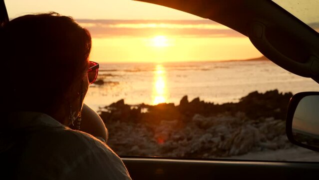 Woman takes in magical vivid sunset whilst sitting in car looking out over ocean