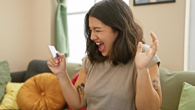 Victorious young hispanic woman ecstatically celebrating a winning achievement at home! sitting on the sofa, credit card in hand, her beautiful smile speaks for her joy and success.