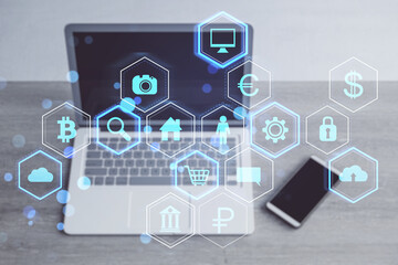 Close up of laptop and mobile phone on wooden desk with abstract hexagonal pattern hologram with various business icons on blurry background. Digital dashboard and data concept. Double exposure.