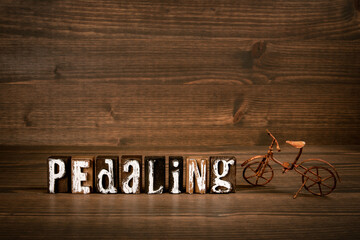 PEDALING. Text from alphabet blocks and rusty miniature bicycle on wood texture background