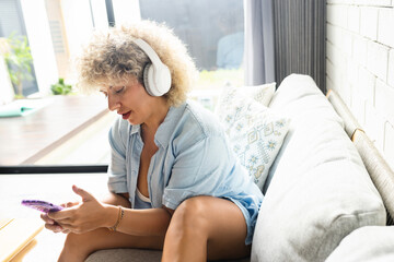 Woman Enjoying Music on Smartphone with Headphones at Home. Relaxed Female Sits Comfortably On A Couch, Engaged In Listening To Music Using Phone And White Headphones,  - 741309094