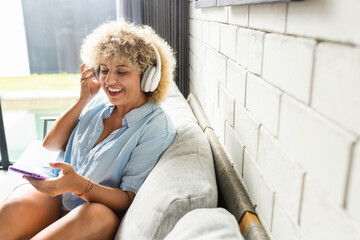 A cheerful woman with curly hair relaxes on a sofa, listening to music using headphones connected...