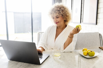 Woman Healthy Morning. A Middle-Aged Female In a White Robe Enjoys a Fresh Apple During Her Morning Routine. Bright Interior, Healthy Lifestyle and Modern Technology  - 741310211