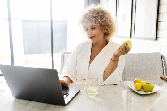 Woman Healthy Morning. A Middle-Aged Female In a White Robe Enjoys a Fresh Apple During Her Morning Routine. Bright Interior, Healthy Lifestyle and Modern Technology 