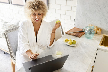 Working From Home. Happy Woman Enjoys a Work-Life Llend at Home, dressed in a comfortable bathrobe while using a laptop and holding a fresh apple. Casual remote work setup in a bright kitchen. - 741310465