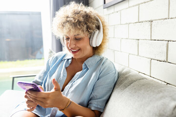 Woman With Headphones Using Smartphone at Home. Young Female Enjoying Music Or Podcast On Her Phone While Relaxing On a Comfortable Couch At Home  - 741310644