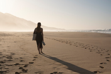 Silueta femenina, bolsa en mano y zapatos en la otra, explorando la playa al atardecer. Libertad,...