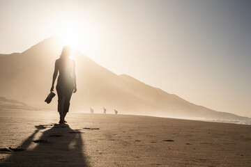 Silueta de una mujer joven disfrutando de una tranquila caminata al atardecer en la suave arena. Serenidad, naturaleza, puesta de sol, bienestar. - obrazy, fototapety, plakaty