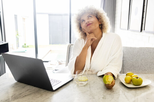 Work From Home. Woman Enjoys a Quiet Moment in a Bathrobe, With a Laptop and a Fresh Apple in Hand. The setting Suggests a Balance of Relaxation, Health.
