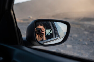 chica sacándose un retrato en el reflejo del espejo retrovisor del coche durante las vacaciones, con una cámara de fotos - obrazy, fototapety, plakaty
