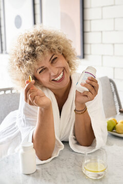 Woman in white bathrobe, laughing and holding a vitamin capsule with a dietary supplement container in hand, in a bright kitchen setting. Promoting health and wellness with a joyful expression.