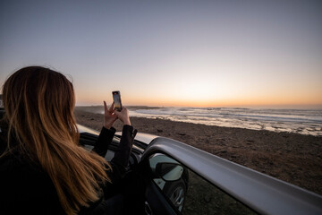 Chica desde su coche de alquiler de vacaciones sacando una foto de la puesta del sol en la playa de Fuerteventura - obrazy, fototapety, plakaty