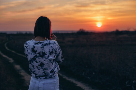 a woman stands in a field with her back to the camera and photographs the sunset on her phone 