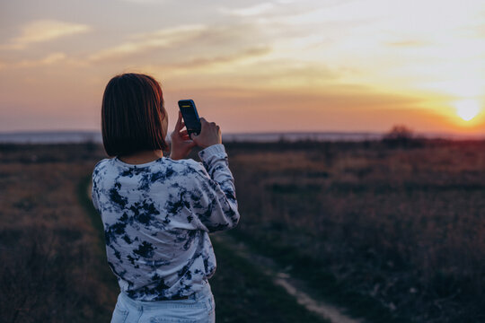 a woman stands in a field with her back to the camera and photographs the sunset on her phone 