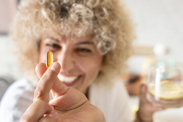 Close-up of a cheerful, curly-haired woman displaying a golden Omega-3 capsule to the camera with a blurry background, symbolizing health, wellness, and dietary supplements at home. - 741312808