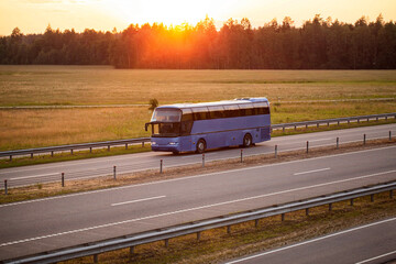 A blue comfortable bus transports passengers along the highway in the evening against the backdrop of sunset. Concept of traveling to another country by bus from a travel agency, convenience © HENADZY